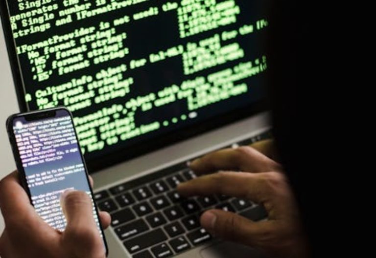 Unrecognisable hacker with smartphone typing on laptop at desk. Image used for illustration. PHOTO/Pexels